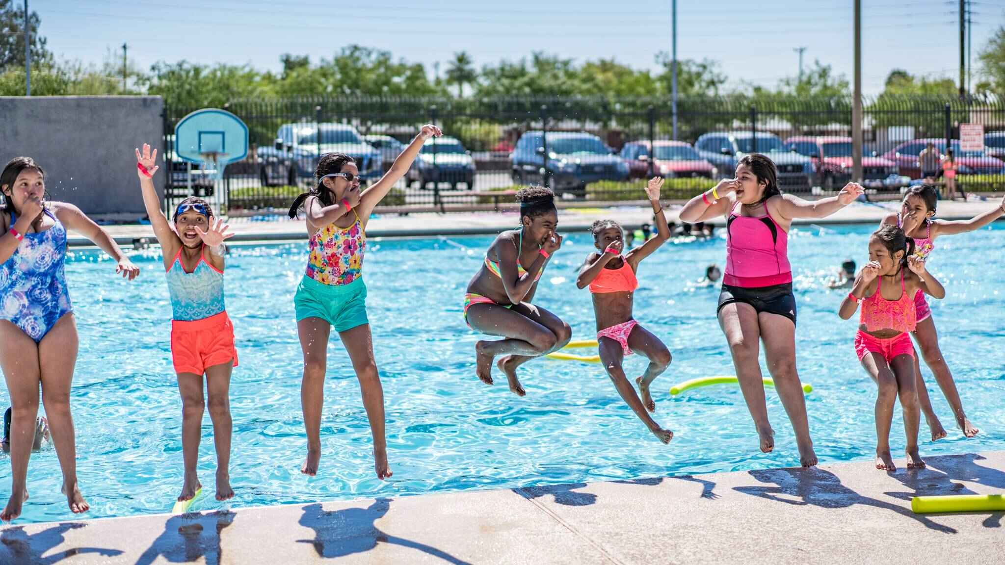 A group of children hold their noses and jump backwards into an outdoor swimming pool.