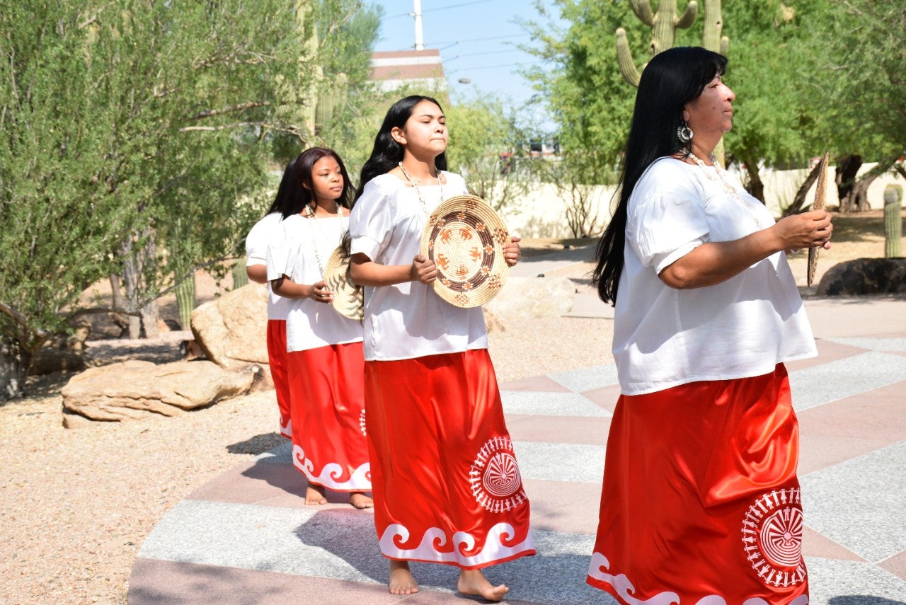 <p>A group of indigenous women move in a procession in an outdoor space. They are wearing white tops and red skirts and are holding woven platters.</p>